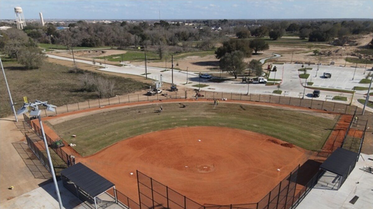 Aerial view of a new red-clay softball field with dugouts and light poles at Primrose Park in Fulshear, with a parking lot, trail and the city water tower in the background.